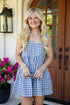A smiling blonde woman stands on a front porch wearing a blue and white gingham sleeveless romper. She is holding sunglasses, with a black lantern, a potted pink flower, and a brown wooden door in the background.