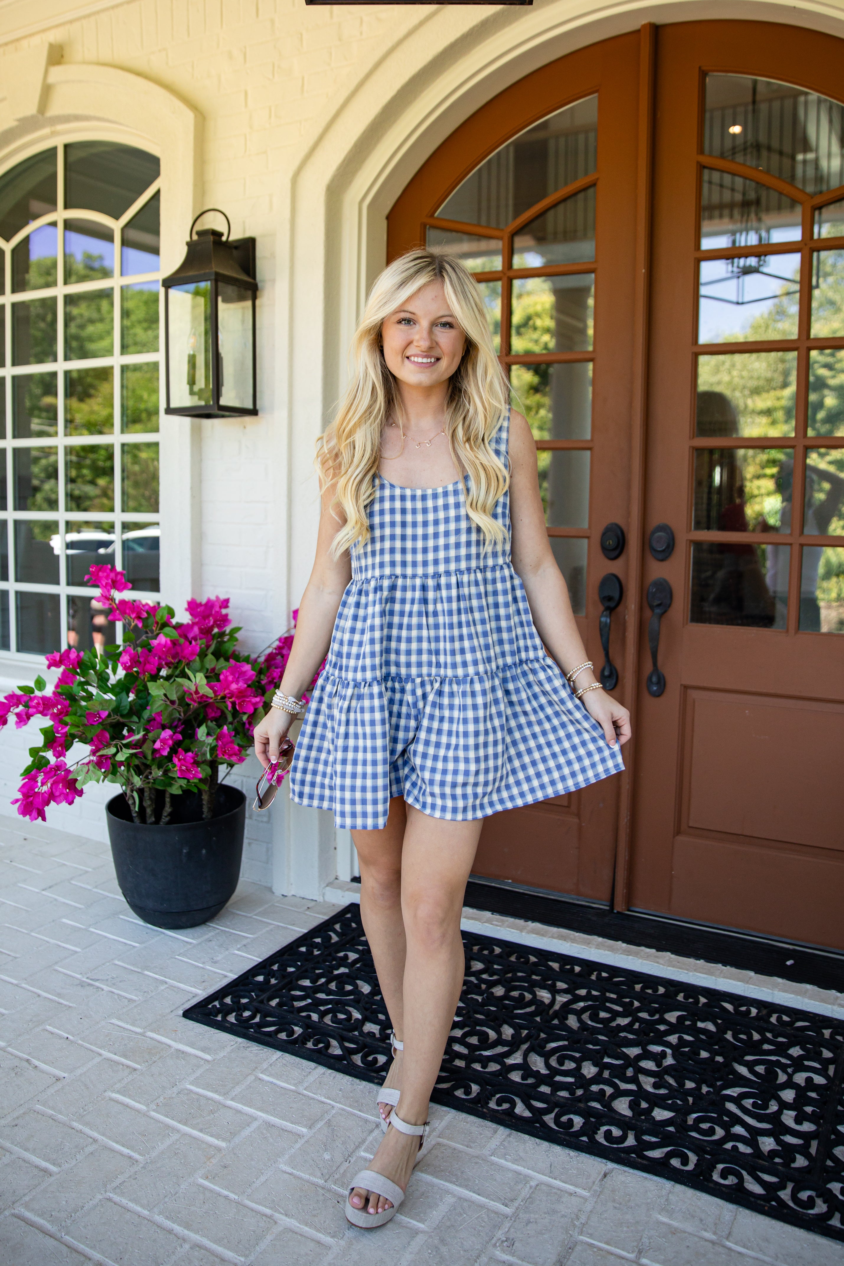 A woman stands on a front porch wearing a blue and white gingham tiered romper dress. The porch features brown double doors, a black doormat, a black lantern, and a potted plant with bright pink flowers.
