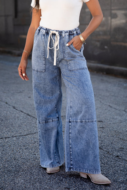 A person wearing light blue wide-leg denim jeans with a white drawstring waist, paired with a white top and beige heeled boots, standing outdoors on asphalt.