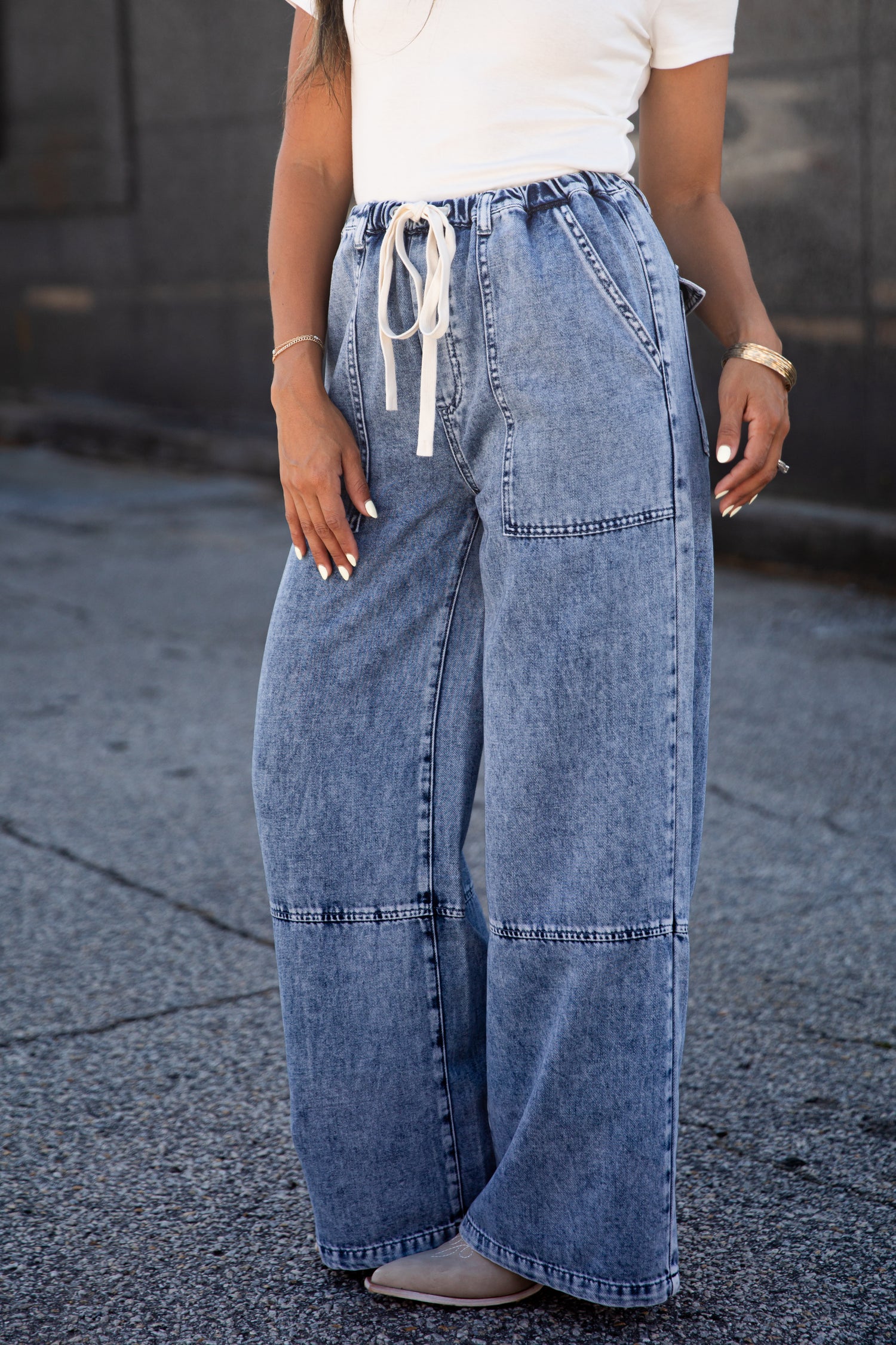 A person wearing wide-leg light blue denim jeans with a white drawstring waist, paired with a white top and gray shoes, standing outdoors on a paved surface.