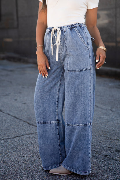 A person wearing wide-leg light blue denim jeans with a white drawstring waist, paired with a white top and gray shoes, standing outdoors on a paved surface.