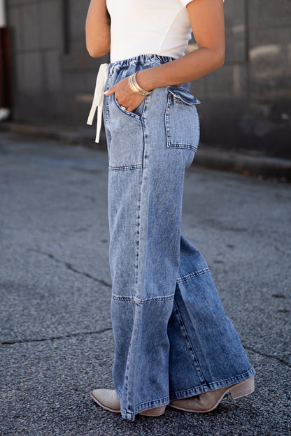 A person wearing wide-leg light blue denim jeans with a white drawstring waist, paired with a white top and beige ankle boots, standing on a paved street.