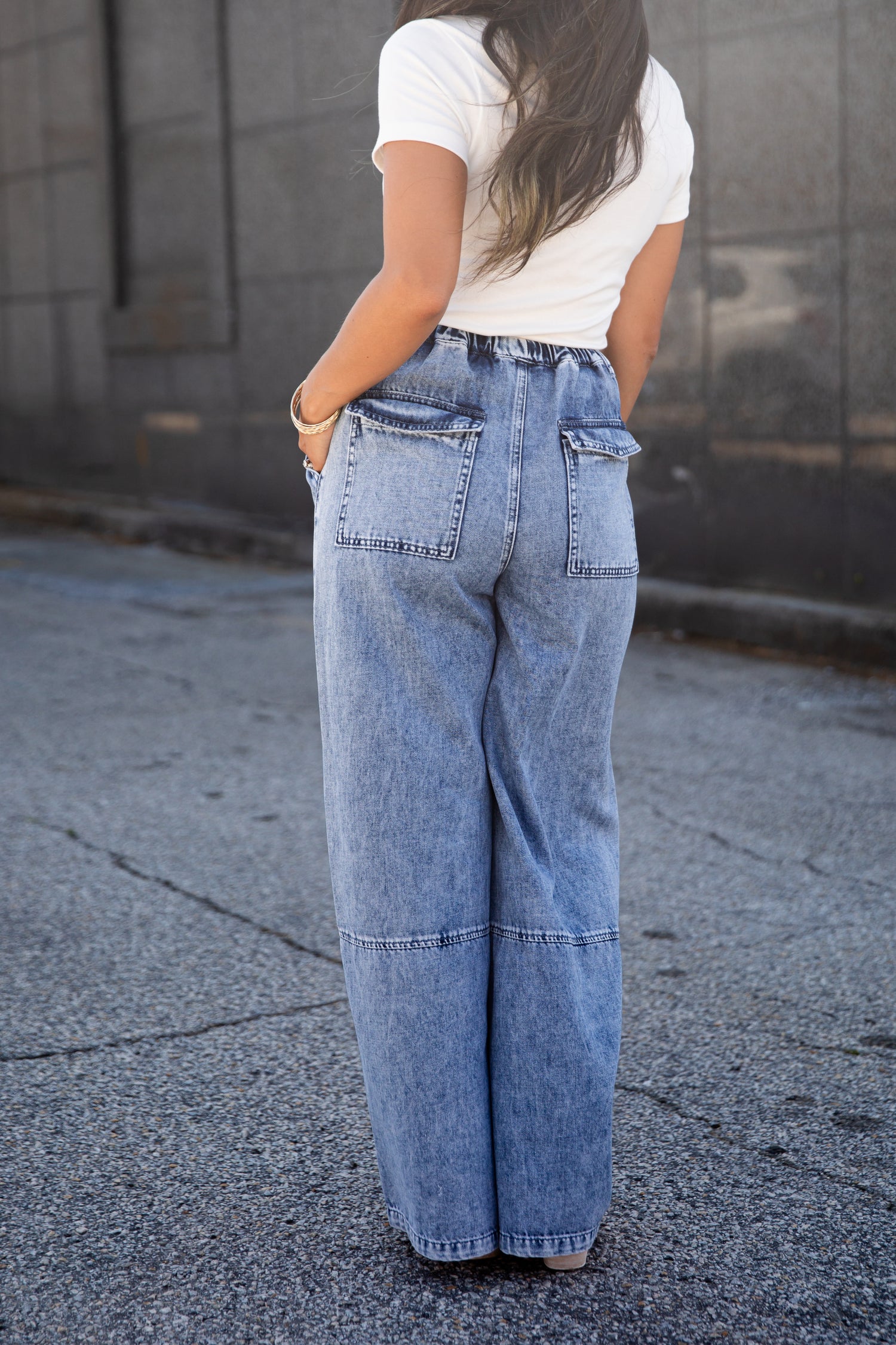 A person viewed from behind wearing light-wash wide-leg denim jeans and a white short-sleeve top, standing outdoors on a paved surface against a gray wall.