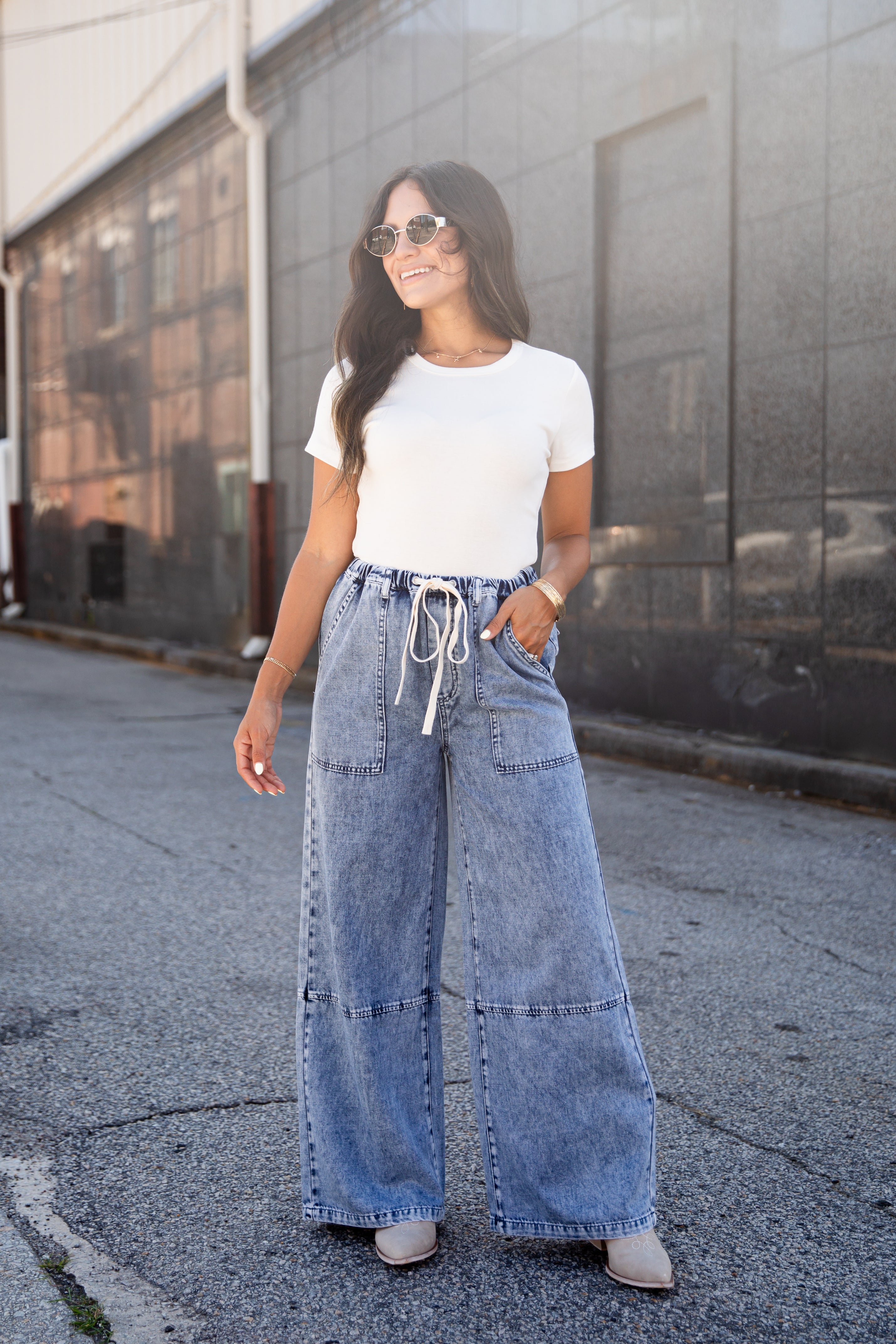 A woman wearing a white short-sleeve crewneck t-shirt and high-waisted, wide-leg light blue denim jeans with a white drawstring waist, standing outdoors on a paved street in front of a gray building.
