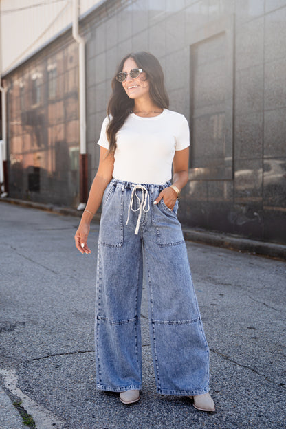 A woman wearing a white short-sleeve crewneck t-shirt and high-waisted, wide-leg light blue denim jeans with a white drawstring waist, standing outdoors on a paved street in front of a gray building.