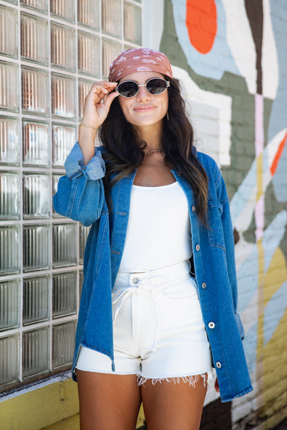 A woman posing outdoors in front of a glass block wall and a colorful mural, wearing a blue denim button-up shirt over a white tank top and white frayed denim shorts, with a pink patterned bandana and sunglasses.