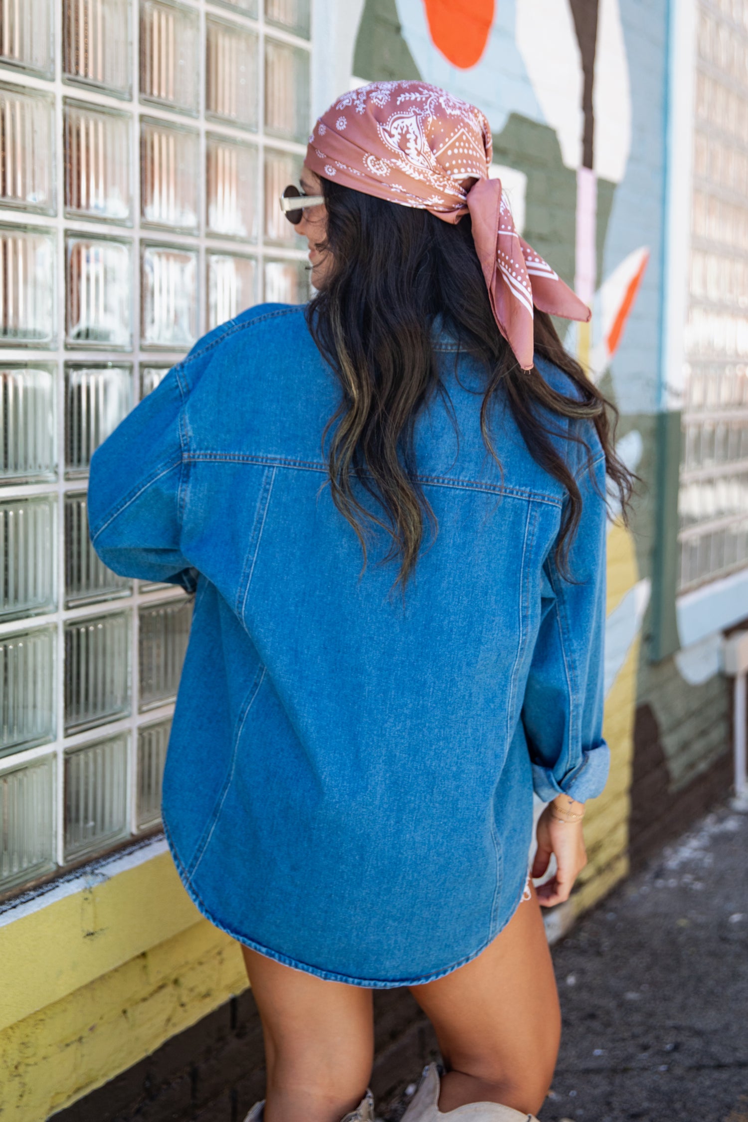 A woman wearing an oversized blue denim shirt and a pink patterned bandana, photographed from behind against a glass block wall and painted wall background.