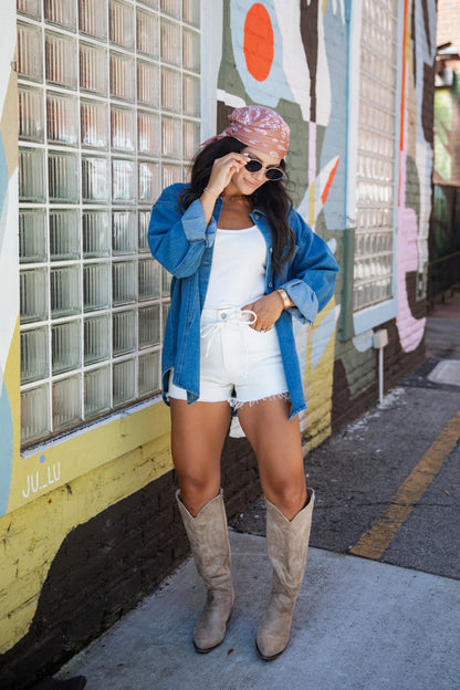 A woman stands outdoors against a painted block-window wall wearing a blue denim shirt, white shorts, beige cowboy boots, and a pink patterned headscarf.