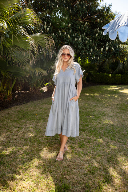 Woman in a light blue dress standing in a garden with trees and grass.