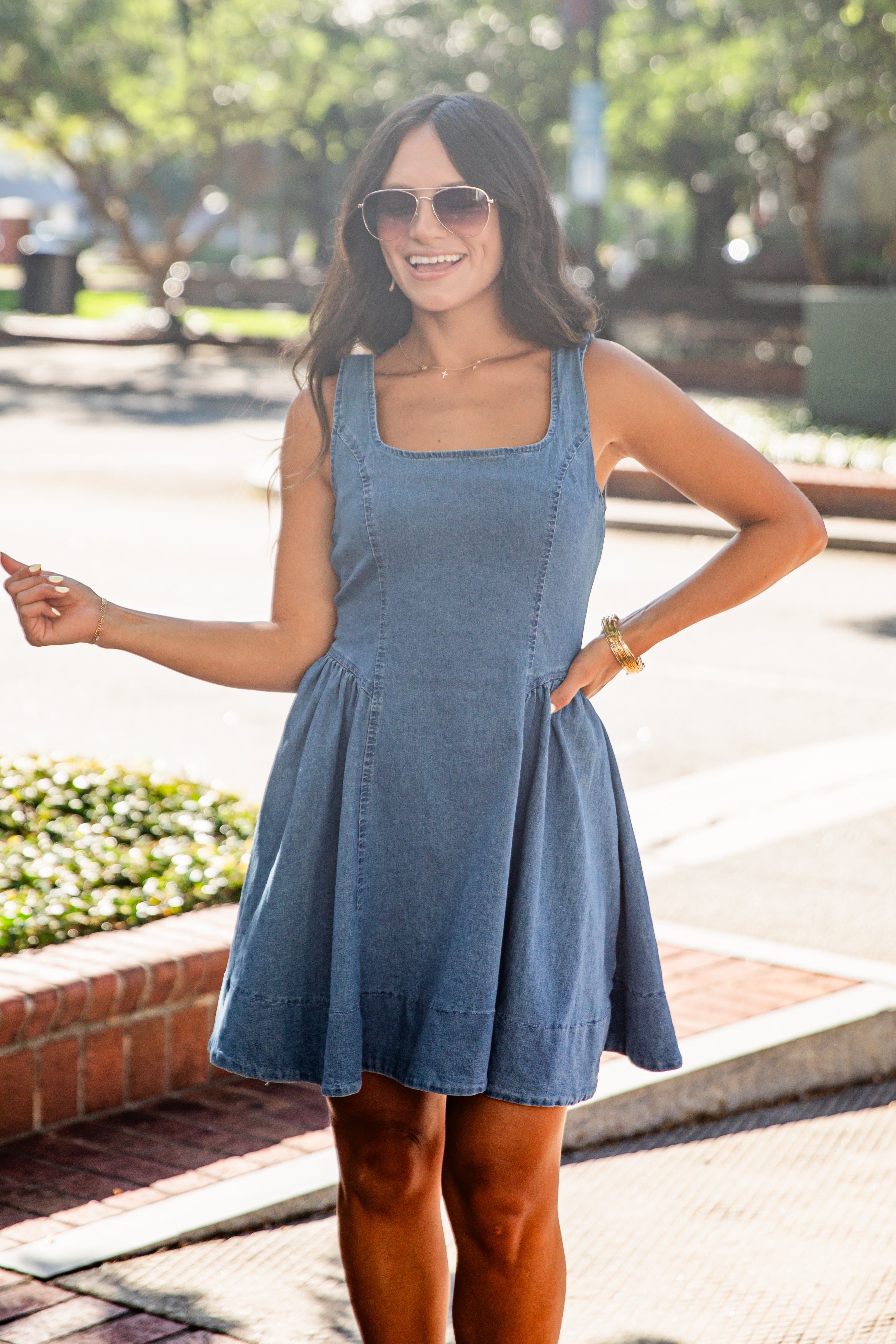 A woman wearing a sleeveless blue denim pinafore dress and sunglasses, standing outdoors on a sidewalk with trees and a street in the background.