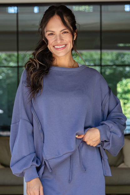 Woman wearing a blue sweatshirt standing in an indoor setting with greenery outside.