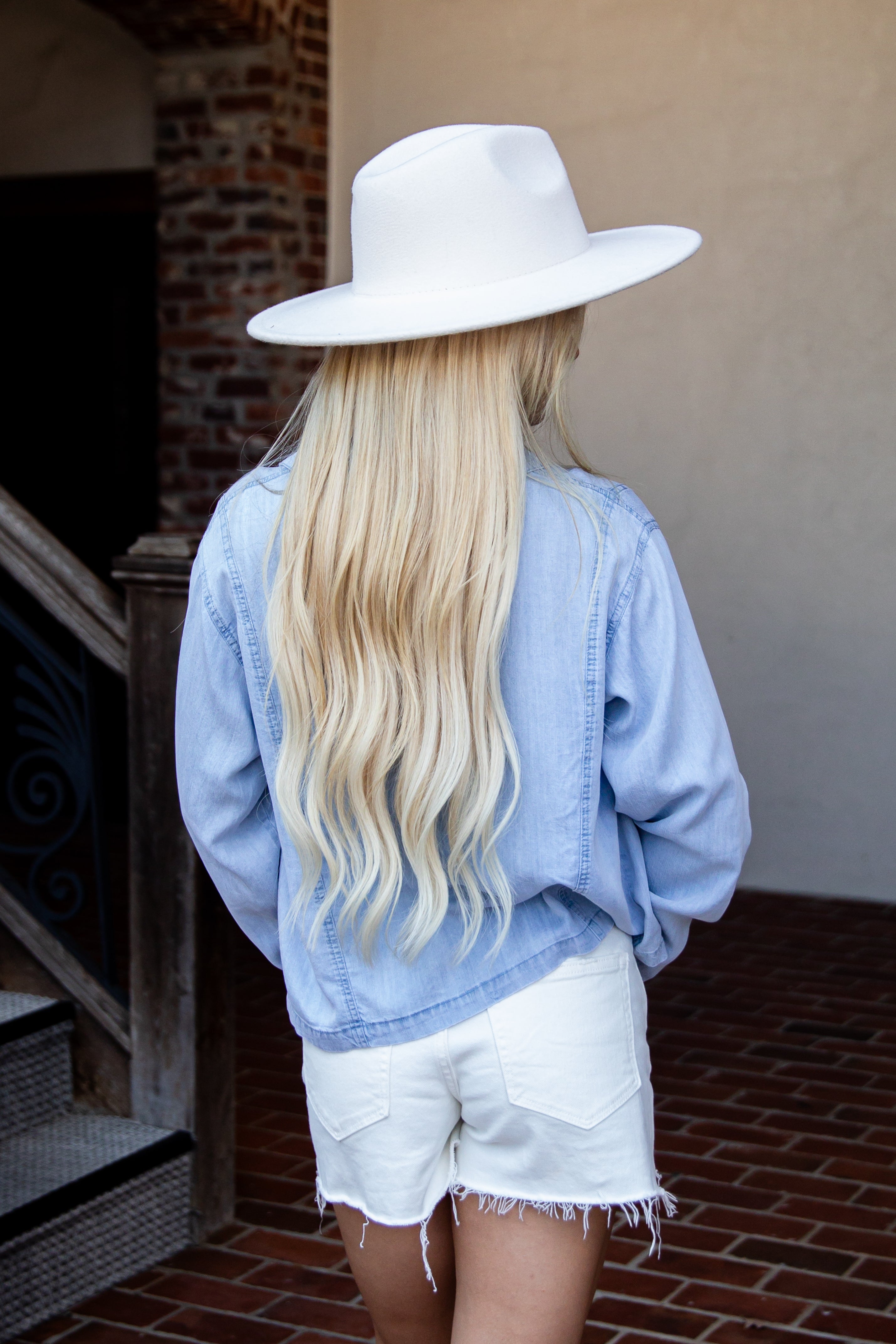 A woman with long blonde hair is shown from behind wearing a white wide-brim hat, a light blue denim shirt, and white denim shorts, standing on a brick patio.