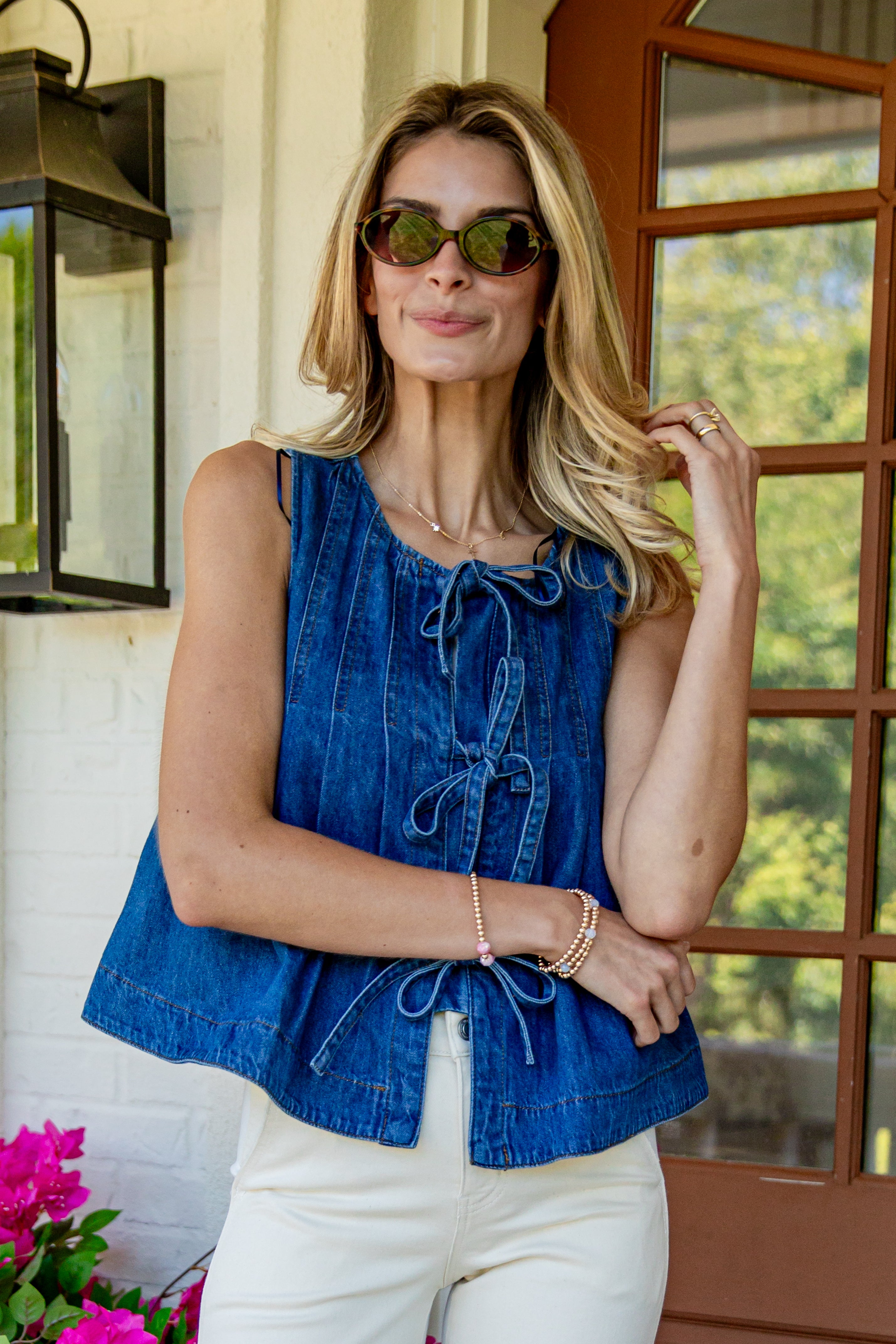 A woman wearing a sleeveless blue denim top with tie-front detailing and white pants, standing on a porch in front of a window and wall lantern.