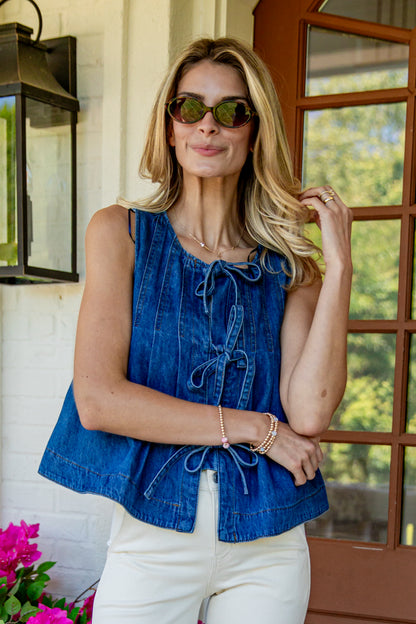 A woman wearing a sleeveless blue denim top with tie-front detailing and white pants, standing on a porch in front of a window and wall lantern.