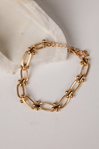 A gold-toned metal chain bracelet with elongated oval links, small bead accents, and a short extender chain, displayed on a white surface next to a white textured block.