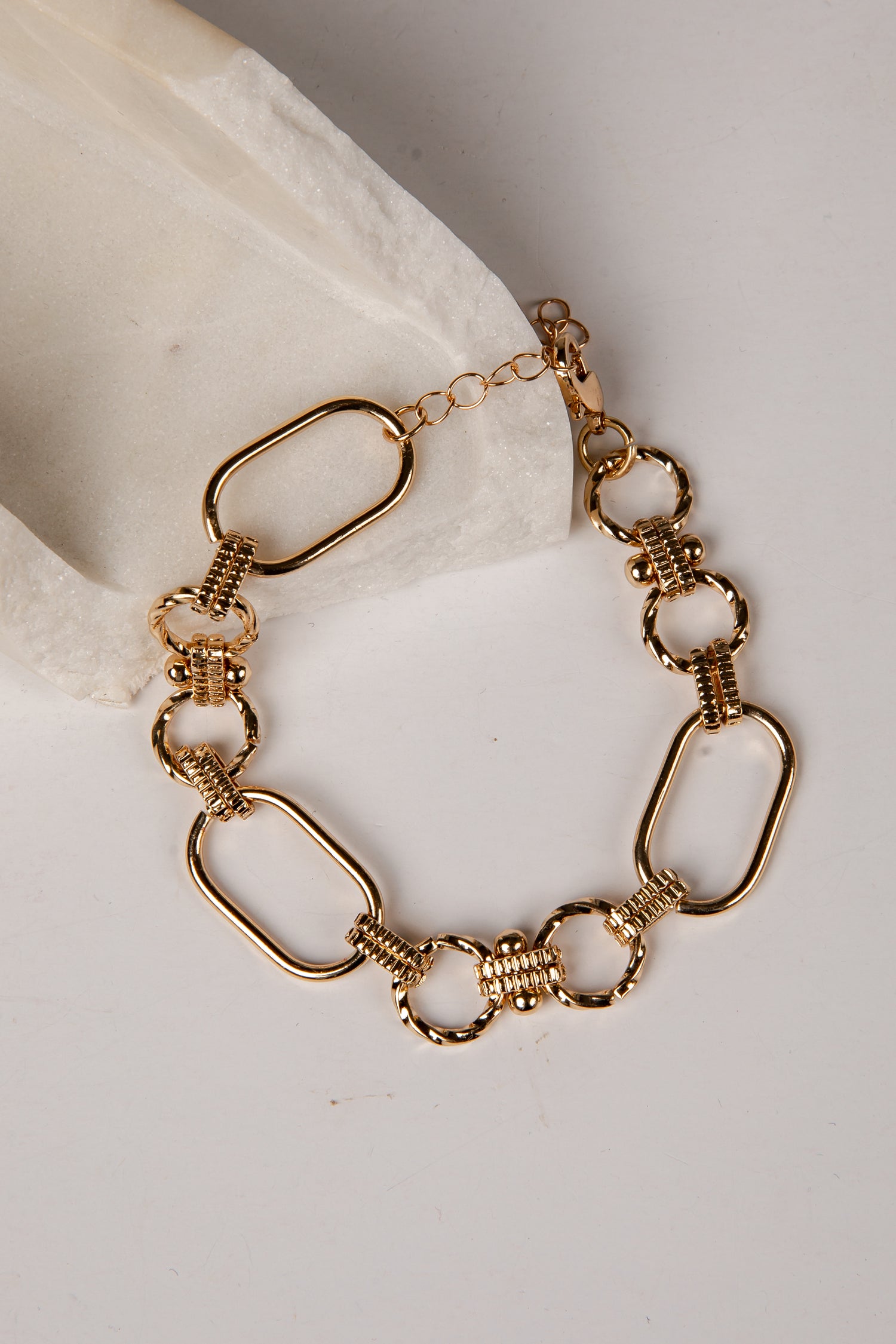 A gold-toned chain bracelet with elongated oval links, textured ring links, and a lobster clasp, displayed on a light neutral surface with a white fabric accent.