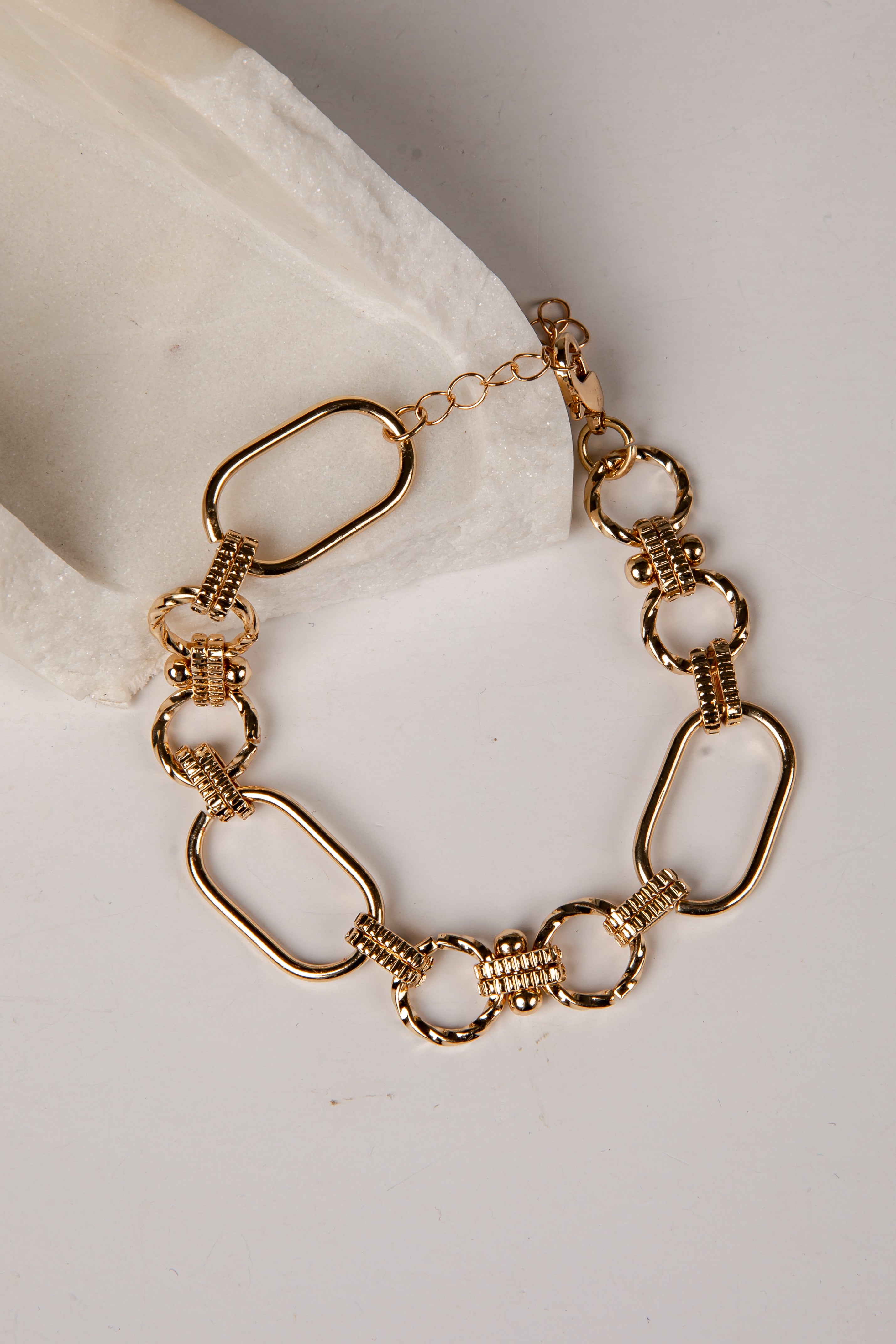 A gold-toned chain bracelet with elongated oval links, textured ring links, and a lobster clasp, displayed on a light neutral surface with a white fabric accent.