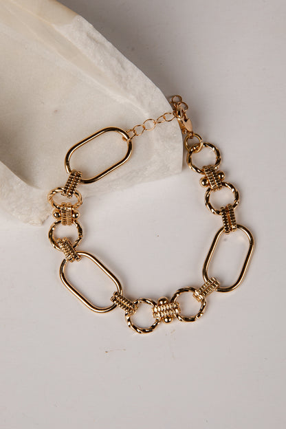A gold-toned chain bracelet with elongated oval links, textured ring links, and a lobster clasp, displayed on a light neutral surface with a white fabric accent.