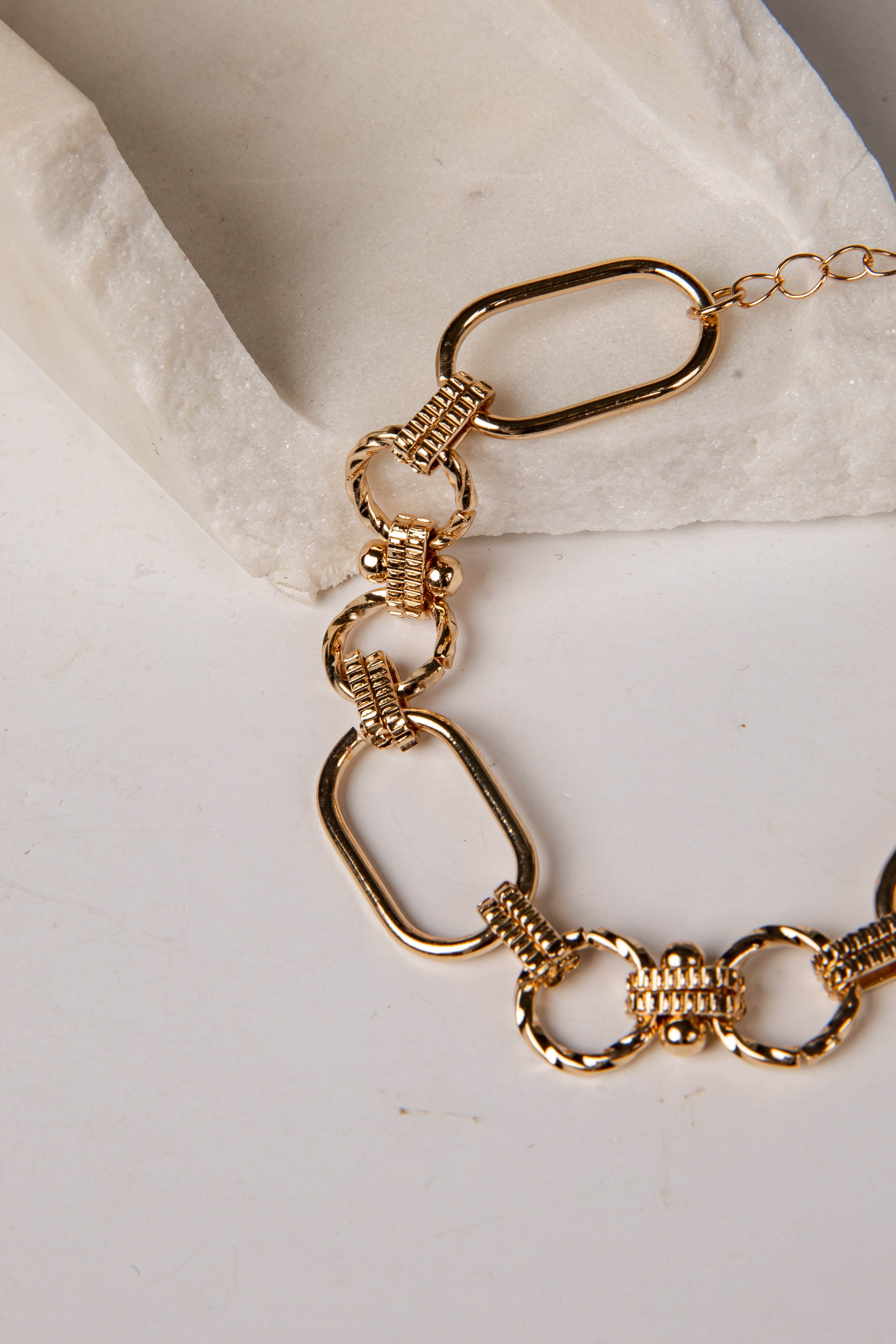 A gold-toned chain link necklace with elongated oval links and textured spacer details, draped over a white textured stone prop on a light surface.