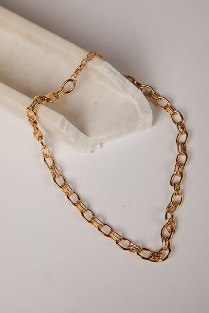 A gold-toned chain necklace with elongated oval links, draped over a white fabric-covered display stand on a light neutral background.