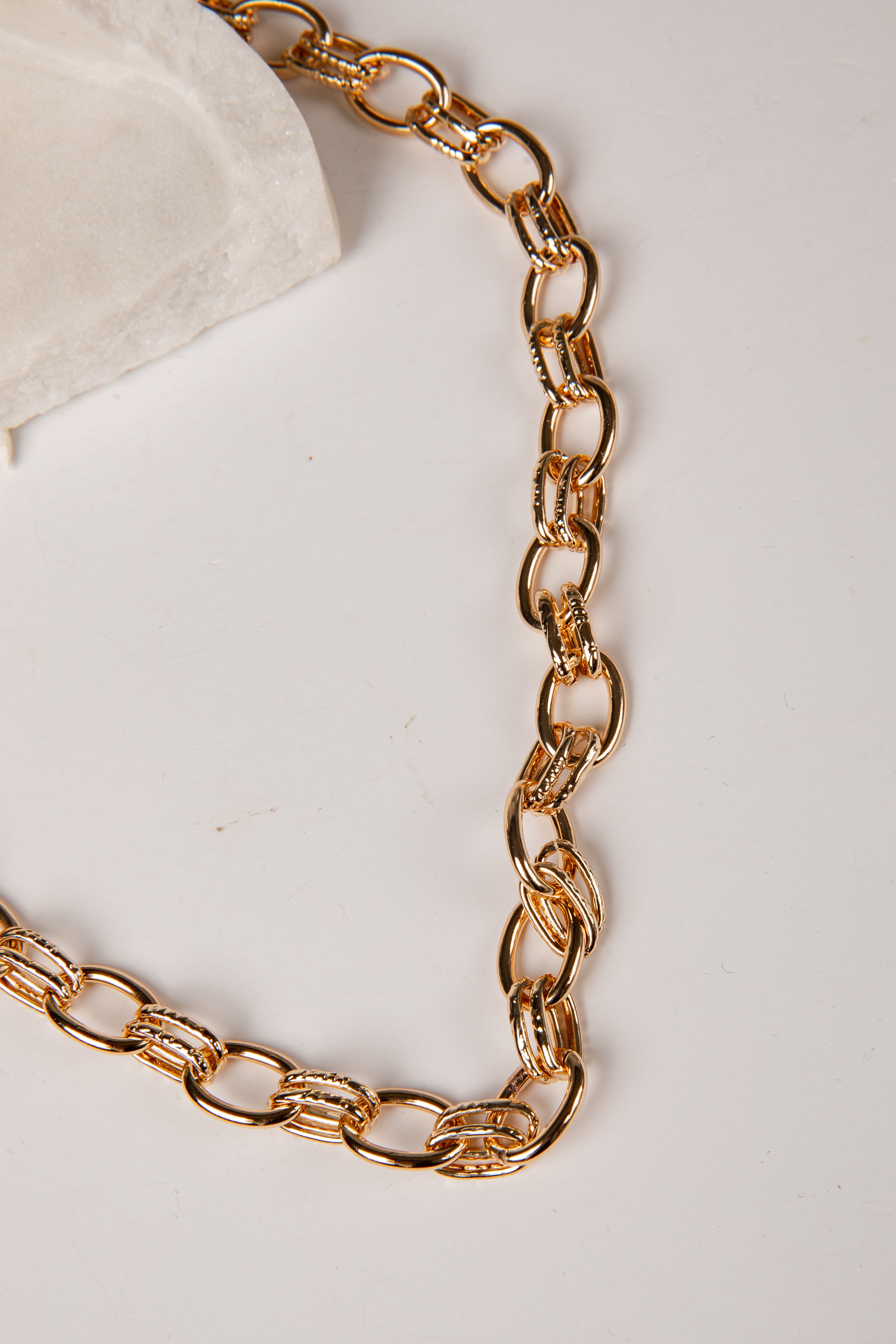 A close-up of a chunky gold chain necklace with interlocking oval links, draped on a light off-white surface with a small white textured prop in the top-left corner.