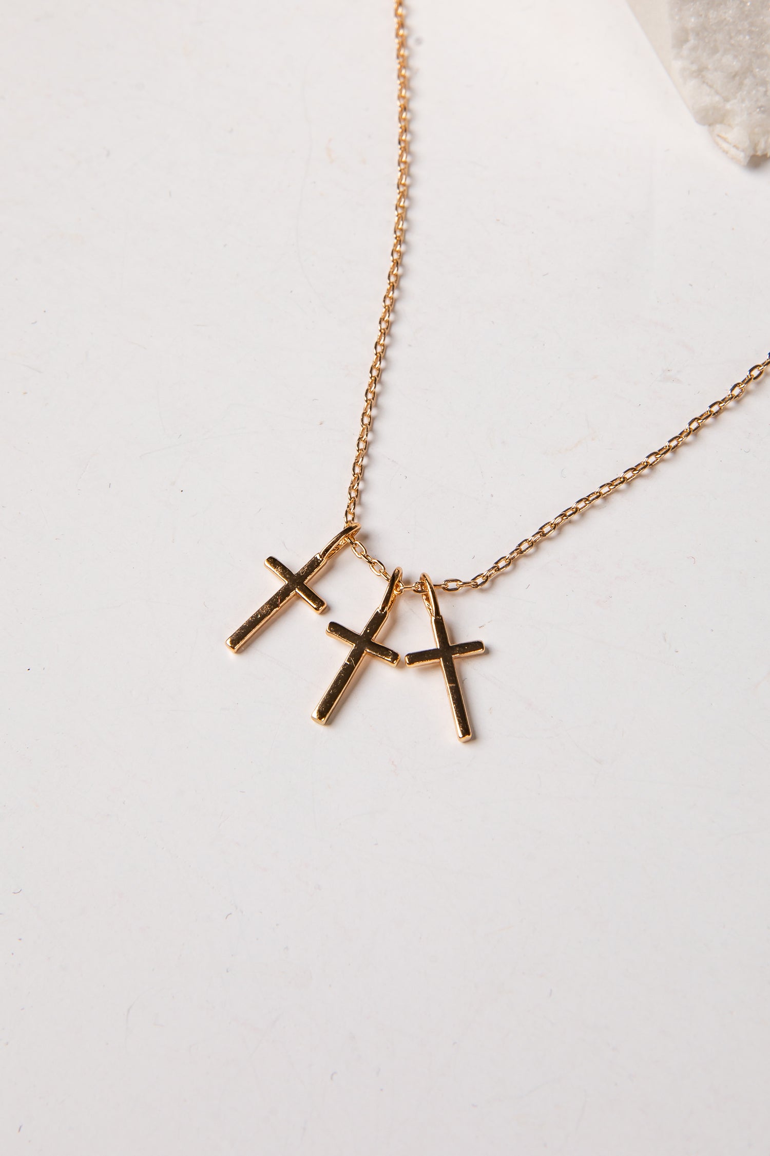 A gold chain necklace with three small cross pendants in a gold-tone finish, displayed on a light off-white surface.