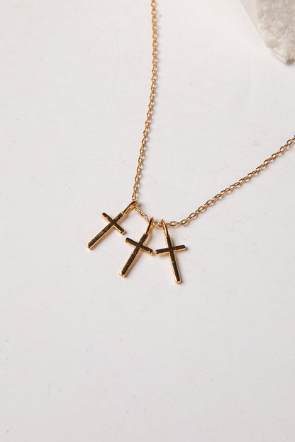 A gold chain necklace with three small cross pendants in a gold-tone finish, displayed on a light off-white surface.