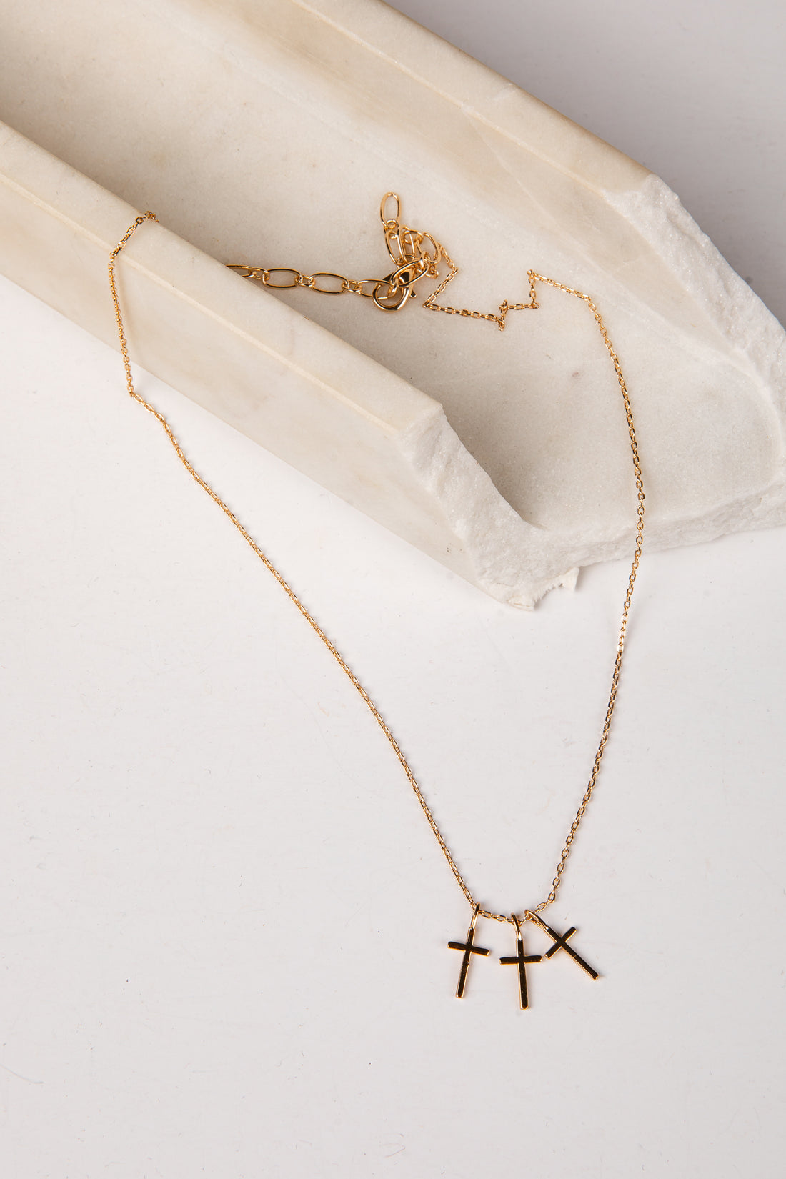 A delicate gold chain necklace with three small cross pendants, displayed on a light cream marble prop against a white background.