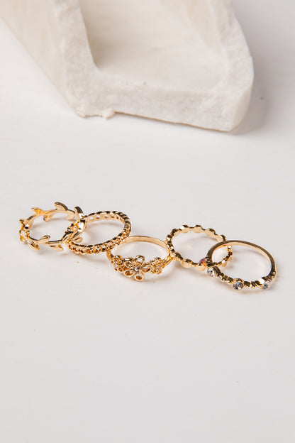 A set of five gold rings with textured, floral, and geometric patterns, arranged in a row on a white surface with a white textured block in the background.