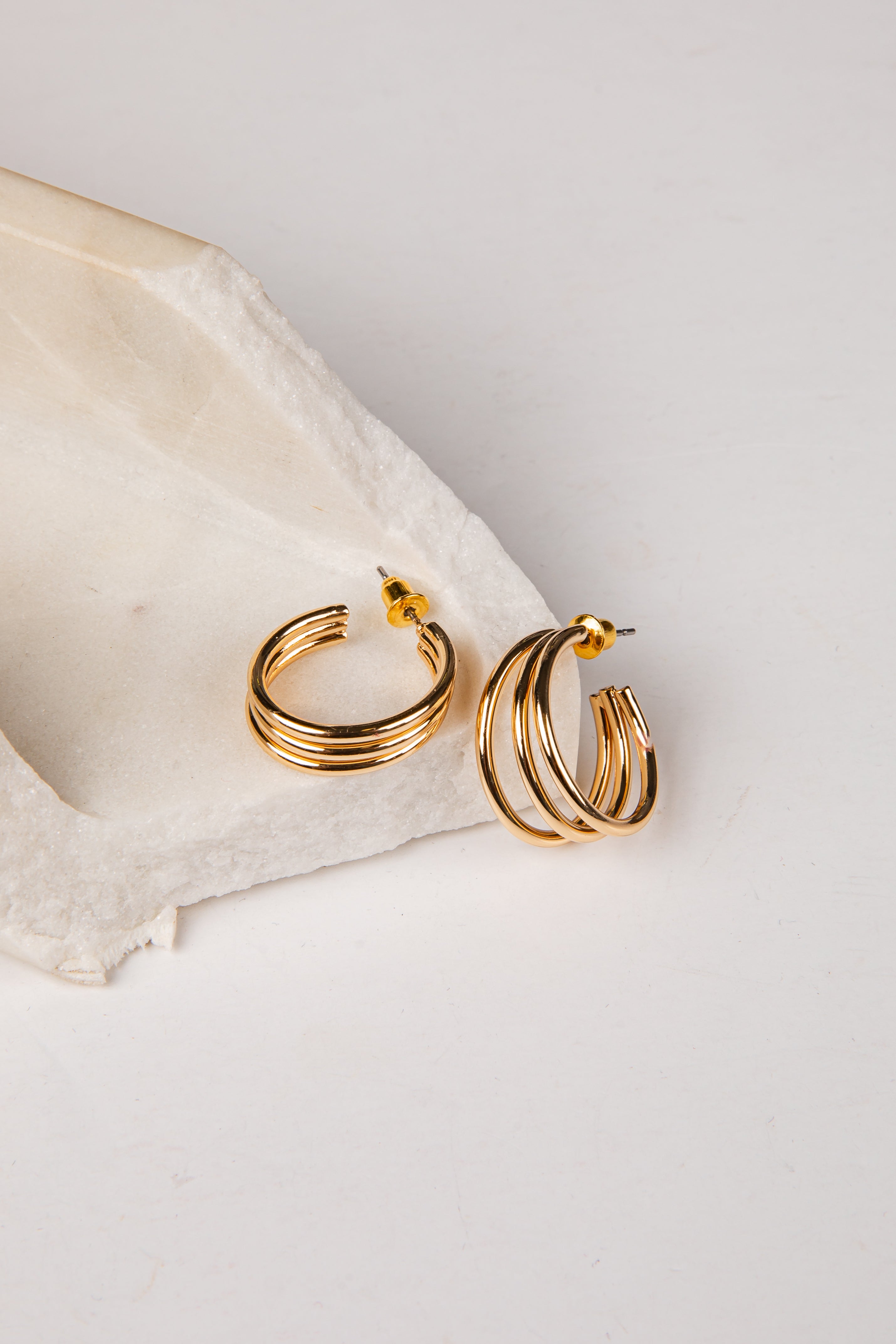 A pair of gold hoop earrings with multiple parallel twisted bands, resting on a textured white stone slab against a clean white background.