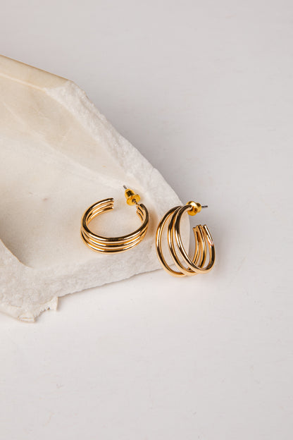 A pair of gold hoop earrings with multiple parallel twisted bands, resting on a textured white stone slab against a clean white background.