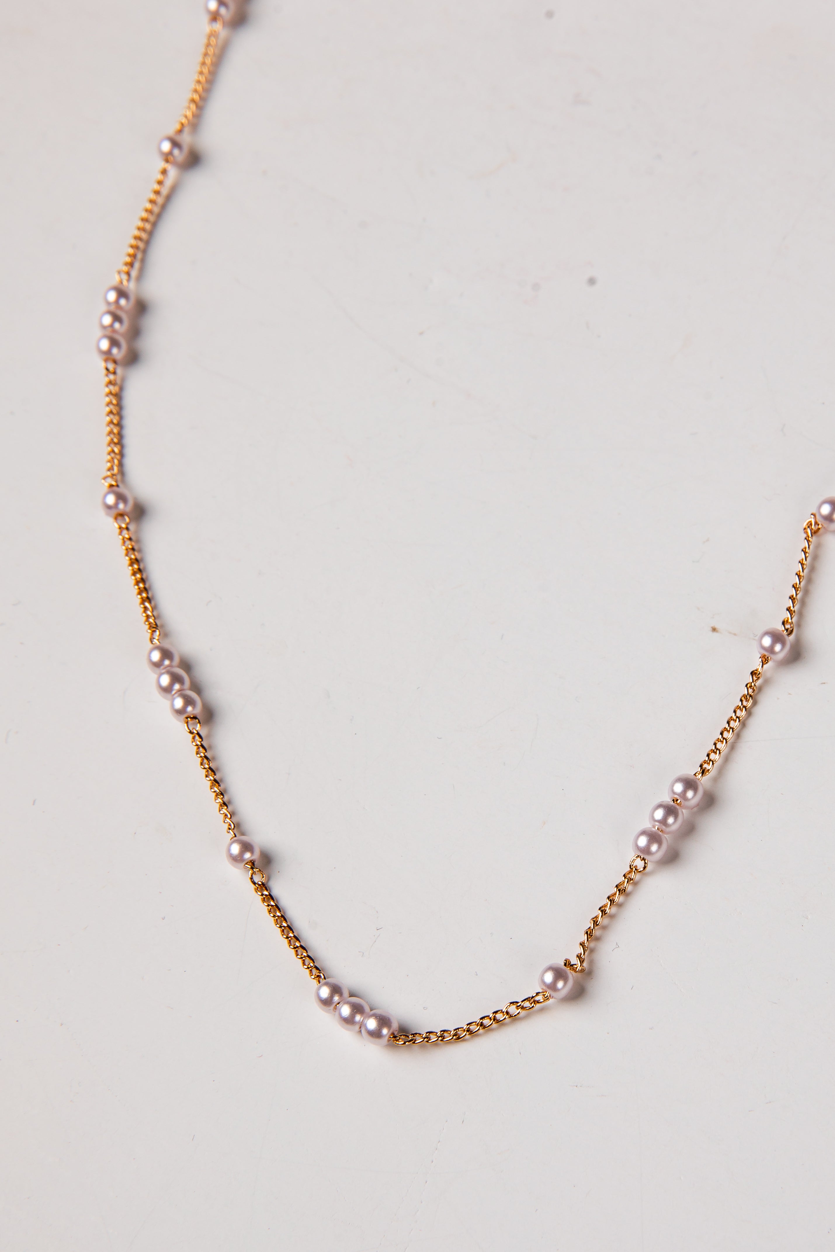 A delicate gold chain necklace featuring small, evenly spaced light pink pearls, laid out on a plain off-white surface.