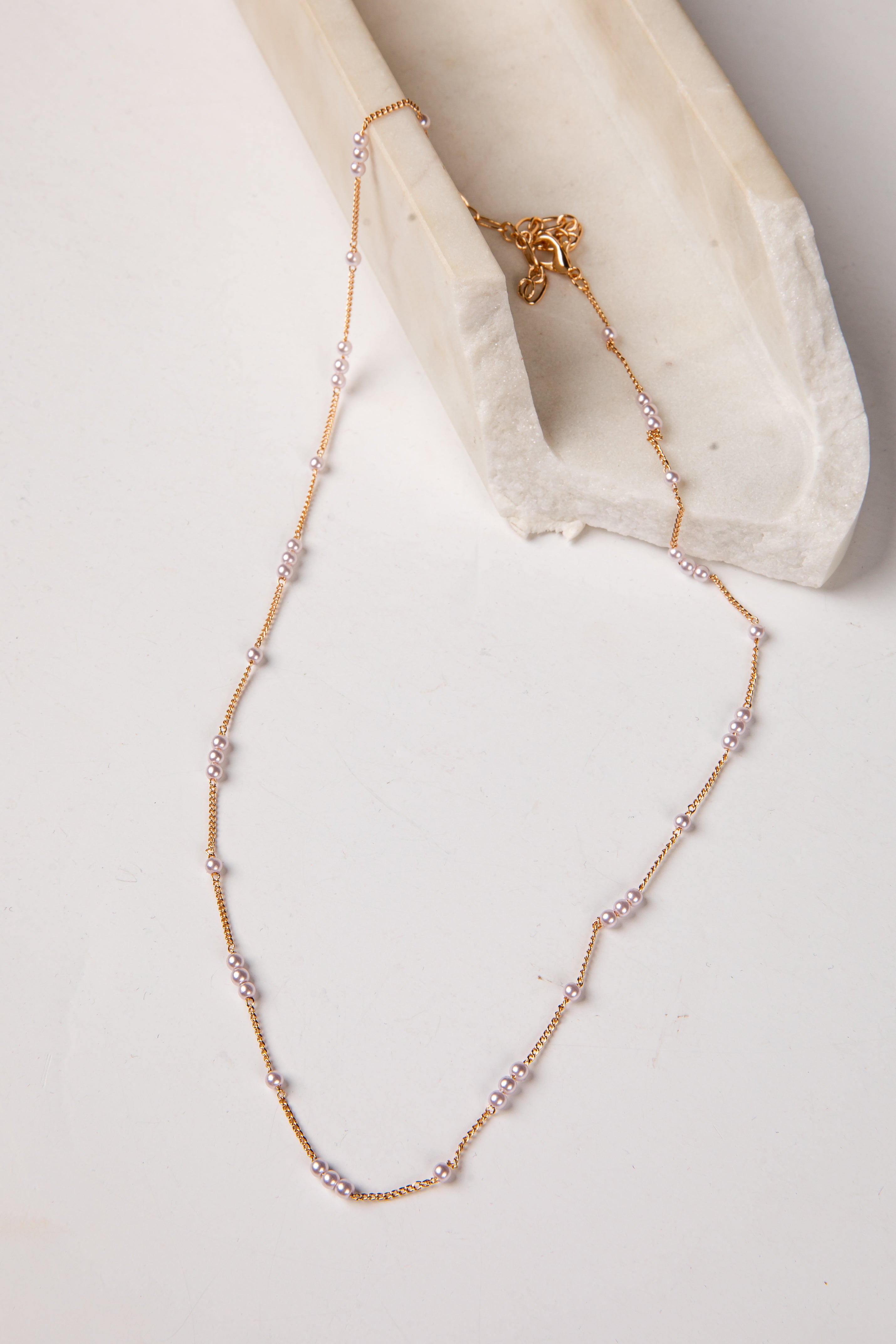 A delicate gold chain necklace with small clear rhinestone clusters spaced along the chain, draped over a white textured fabric prop on a light neutral background.