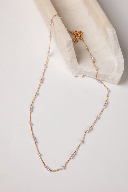 A delicate gold chain necklace with small clear rhinestone clusters spaced along the chain, draped over a white textured fabric prop on a light neutral background.