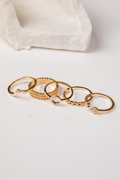 Five gold-toned rings with various textured and pave designs, arranged in a row on a white surface with a soft-focus white textured prop in the background.