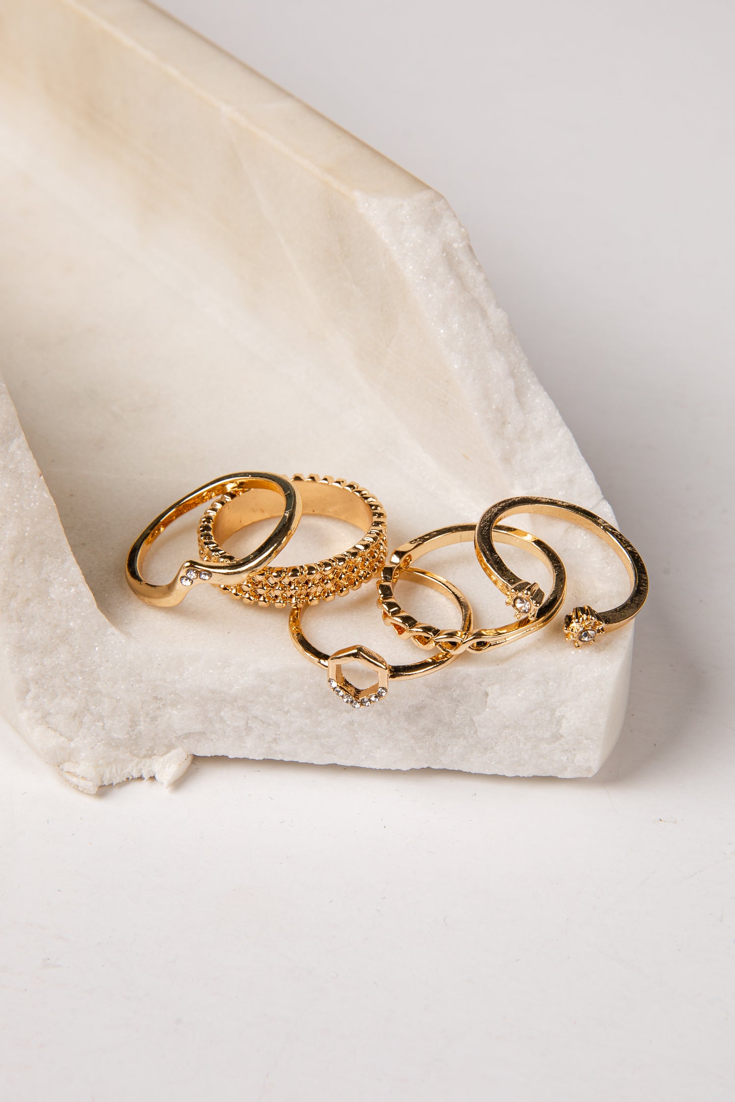 A set of five gold rings with various textures and designs, including a heart-shaped ring and a beaded ring, displayed on a white textured stone tray against a light gray background.