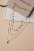 Gold necklace with green stones on a beige surface with a Christian Dior book in the background