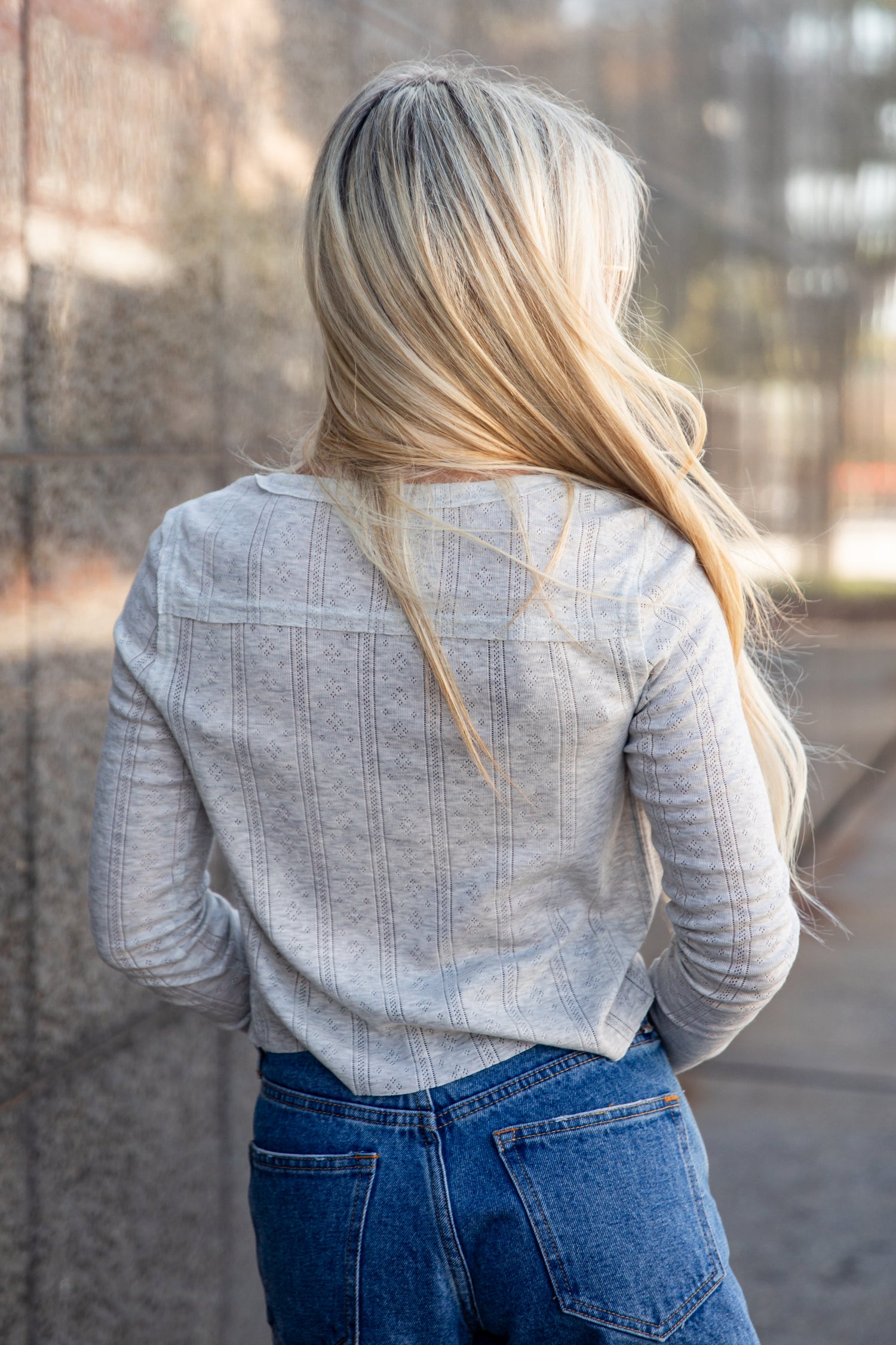 A woman viewed from behind wearing a light gray ribbed long-sleeve top and blue jeans, standing outdoors against a blurred stone wall.