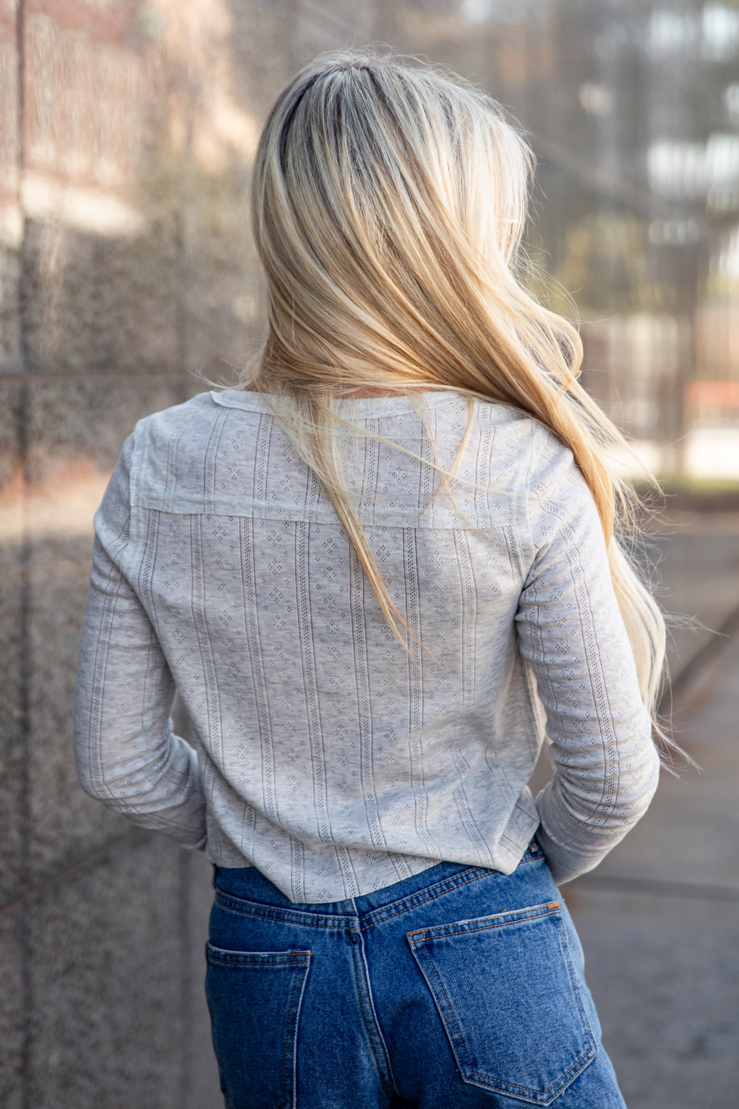 A woman viewed from behind wearing a light gray ribbed long-sleeve top and blue jeans, standing outdoors against a blurred stone wall.