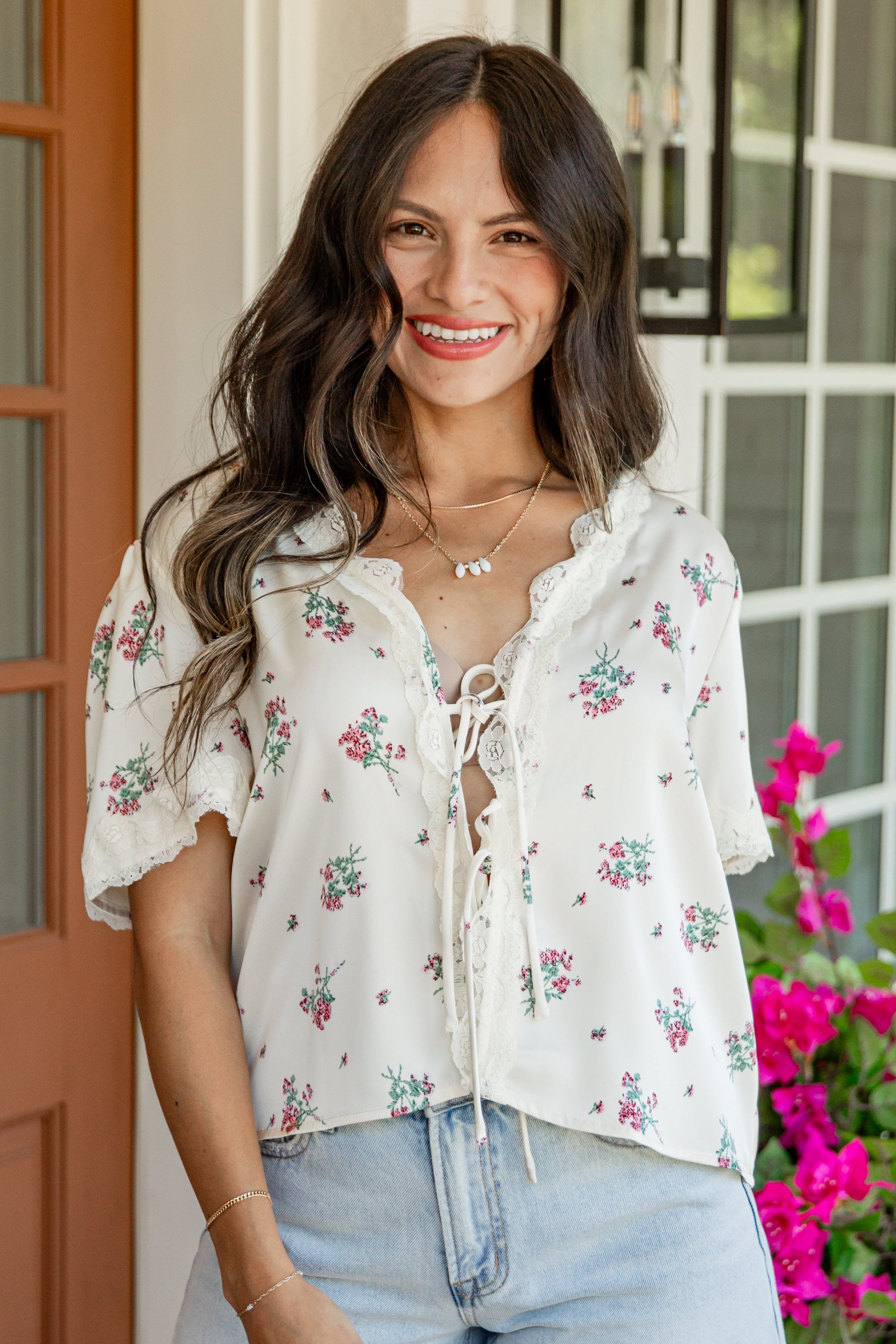 A woman smiling while wearing a white short-sleeve blouse with a pink and green floral print, ruffled collar, and front tie closures, paired with light-wash denim shorts in an outdoor porch setting.