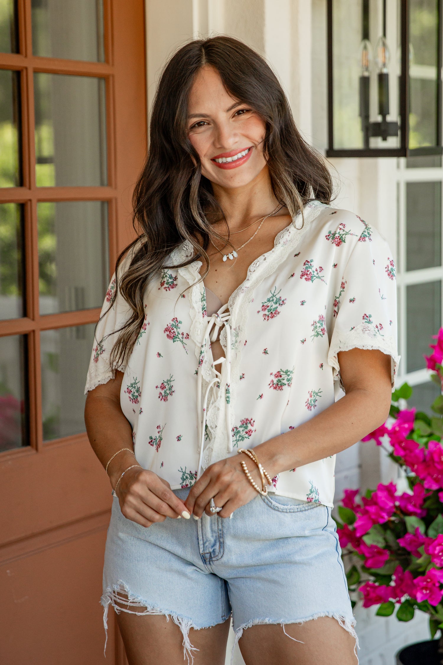 A woman smiling while wearing a short-sleeve white blouse with a delicate pink and green floral print, paired with light-wash denim shorts, standing on a porch.