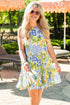 Woman wearing a blue and white tropical print mini dress featuring a sleeveless design, high neckline, and tiered skirt. The vibrant mix of floral, citrus, and palm motifs creates a playful resort-inspired look perfect for vacations, brunches, and sunny summer days.