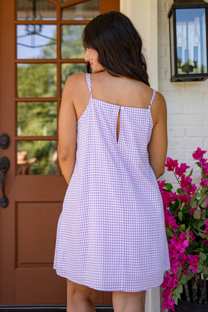 Woman wearing a checkered dress standing in front of a door with flowers to her right.