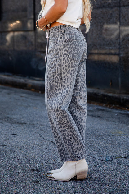 A person wearing loose, wide-leg gray leopard-print jeans with a white cropped top and white ankle boots, standing outdoors on a paved street against a dark stone wall.