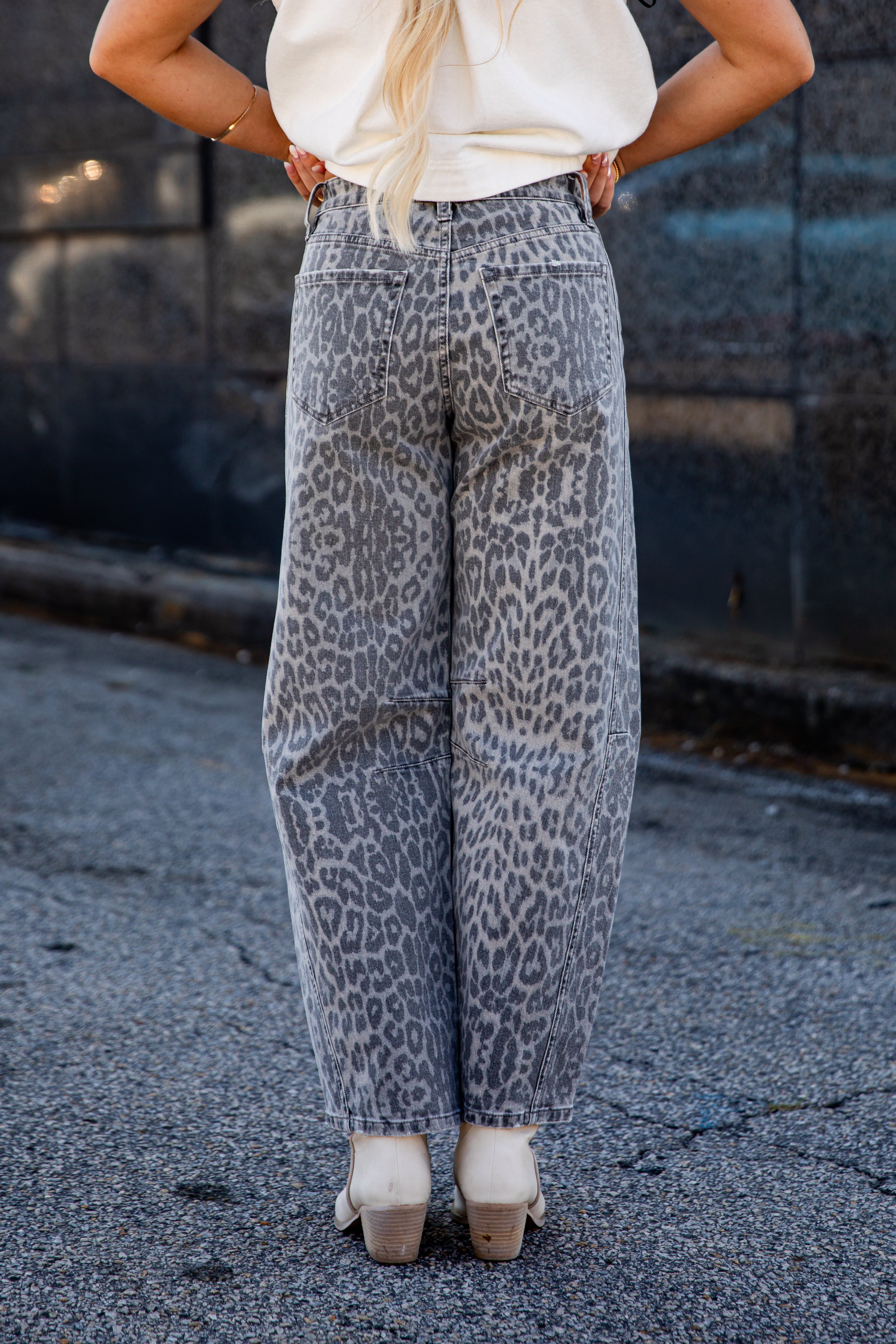 A person shown from the back wearing wide-leg gray pants with a black leopard print pattern, paired with a cream top and beige heels, standing on asphalt.