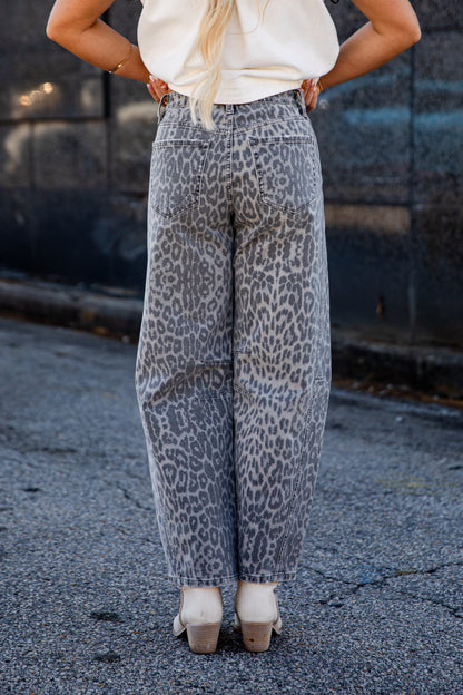 A person shown from the back wearing wide-leg gray pants with a black leopard print pattern, paired with a cream top and beige heels, standing on asphalt.