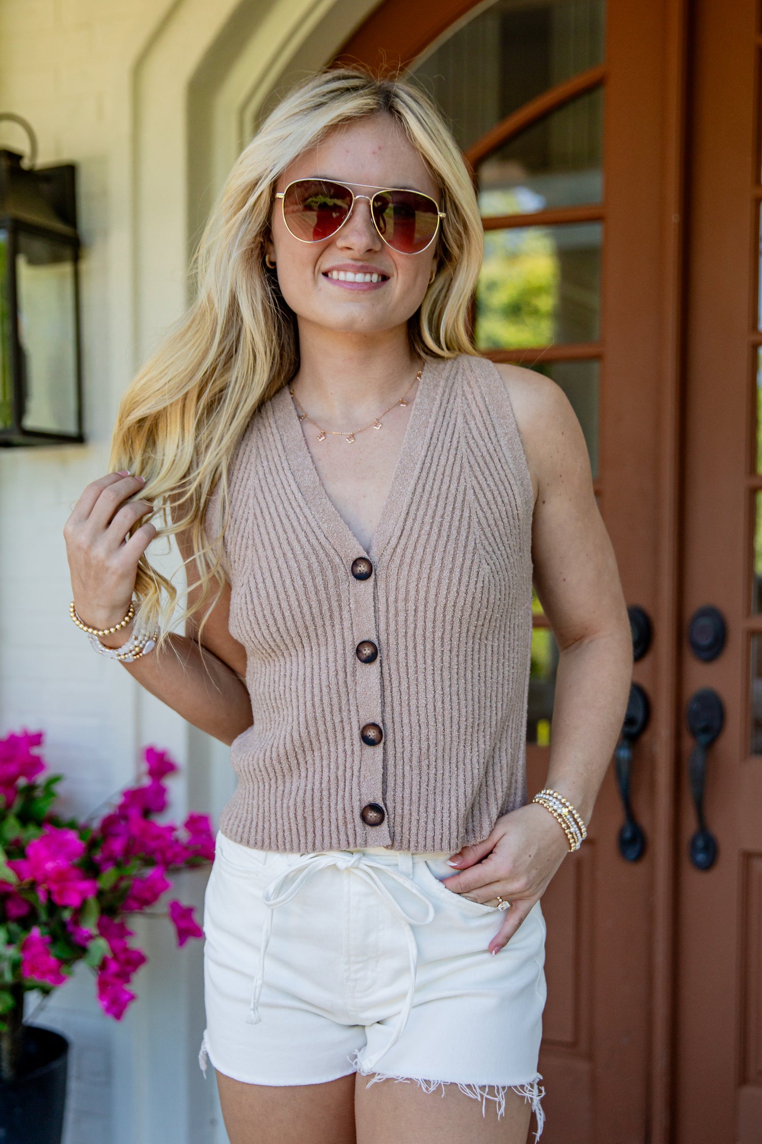 A woman wearing a sleeveless ribbed knit button-front vest in a warm taupe-beige color, paired with white shorts and sunglasses, standing in front of a brown wooden door.