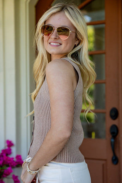 A woman with blonde hair wearing a beige ribbed knit sleeveless top, white pants, and gold-framed sunglasses, standing in front of a brown wooden door.