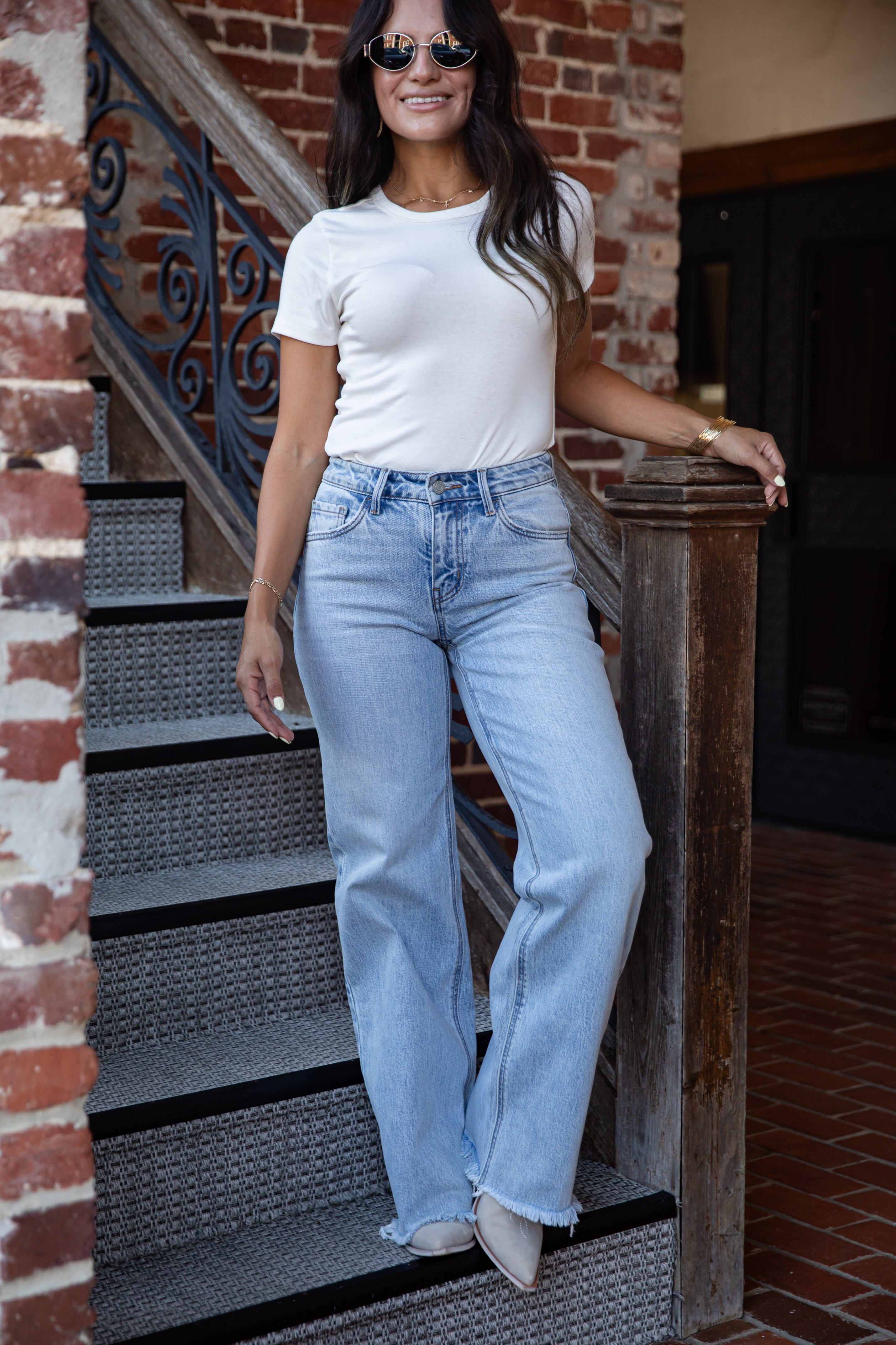 A woman wearing a white short-sleeve crewneck t-shirt and light-wash high-rise flare jeans, standing on outdoor brick stairs next to a wooden railing.