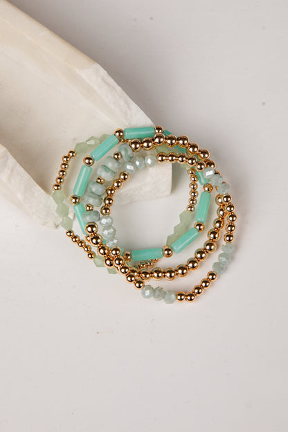 A stack of beaded stretch bracelets featuring light aqua green beads and metallic gold beads, displayed on a neutral cream-colored surface.